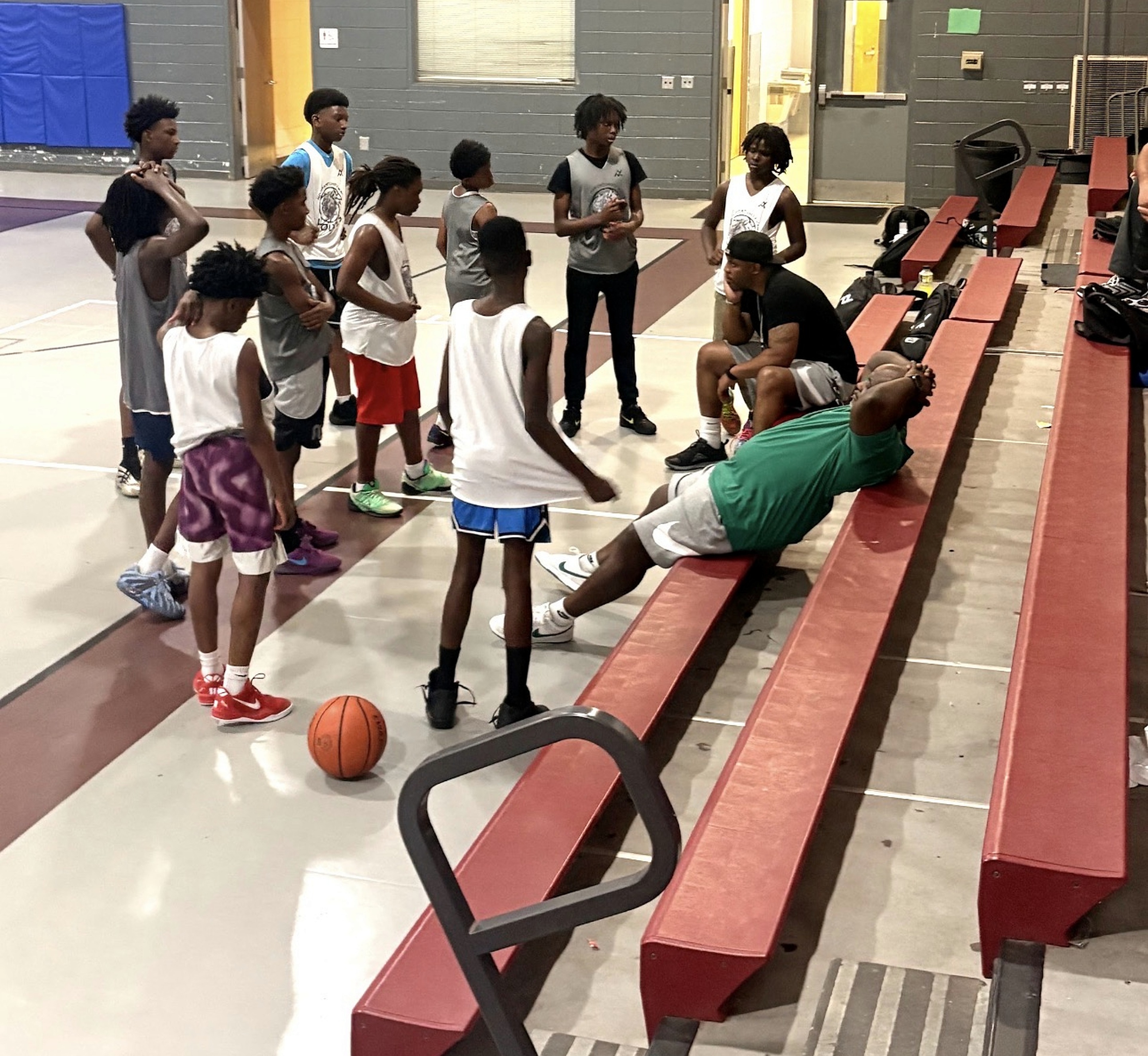 Players listening beside red bleachers with basketballs on the gym floor
